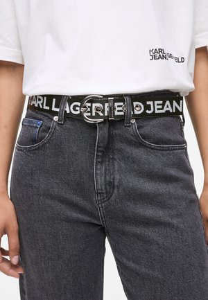 Schwarzer geflochtener Gürtel mit einer runden silbernen Schnalle, auf der der eingestochene Text "KARL LAGERFELD JEANS" in weißer Schrift zu sehen ist. Getragen zu grauen Jeans.