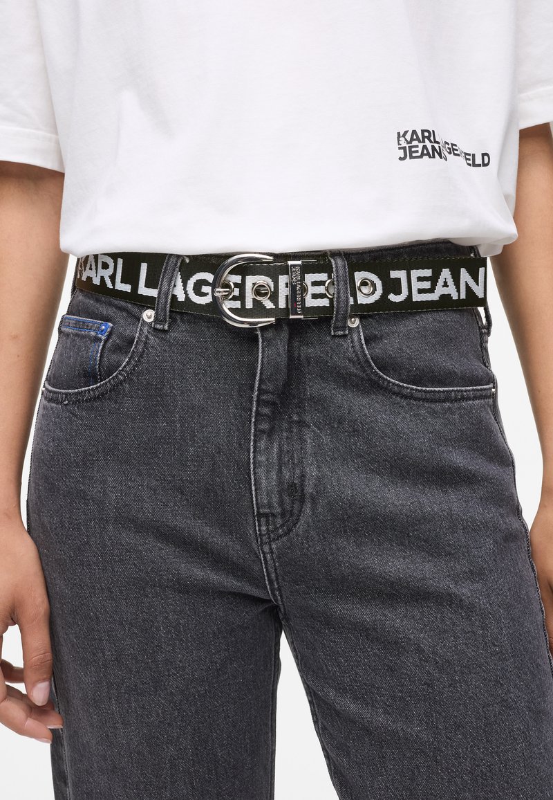 Cintura intrecciata nera con fibbia circolare in argento, caratterizzata dal testo impresso "KARL LAGERFELD JEANS" in lettere bianche. Indossata con jeans grigi.