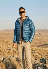 Man wearing blue puffer jacket, beige cargo pants, dark shirt, and large black sunglasses standing on rocky terrain with dry grass and hills in background.