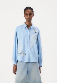 Camisa riscada de azul claro com mangas longas, designs florais bordados, fecho de botão e um corte descontraído, combinada com uma saia de ganga.
