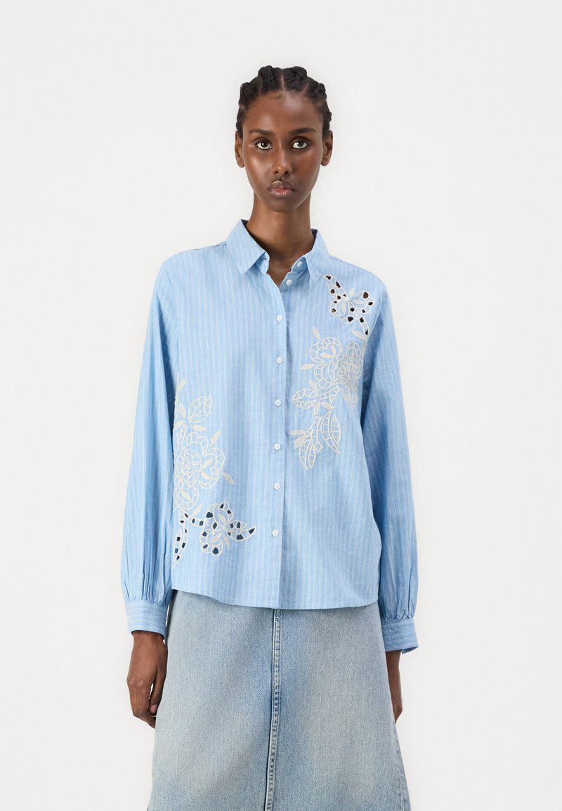 Camisa riscada de azul claro com mangas longas, designs florais bordados, fecho de botão e um corte descontraído, combinada com uma saia de ganga.