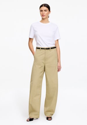 Kobieta stojąca w prostym białym t-shircie z krótkim rękawem, beżowych szerokich spodniach z czarnym paskiem oraz czarnych sandałach na obcasie z odkrytymi palcami.