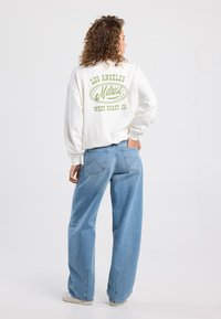 White fleece sweatshirt with green graphic text “Los Angeles Melrose West Coast, CA” on the back, paired with loose blue denim jeans.