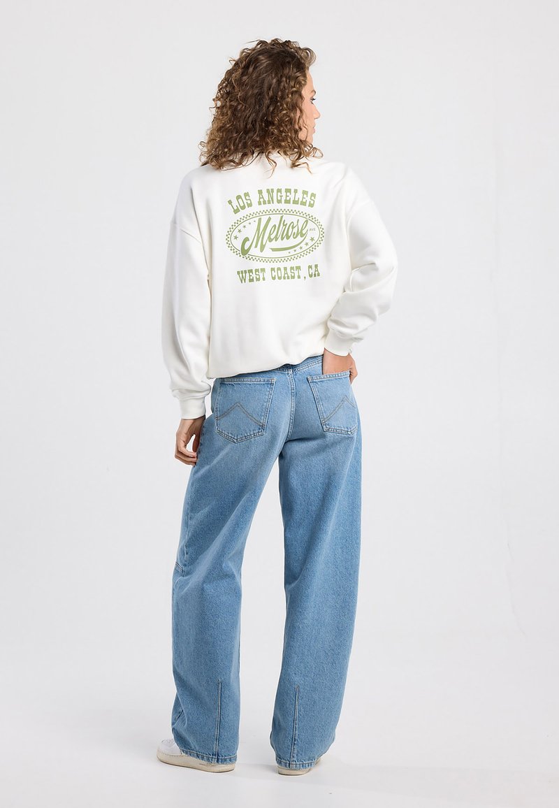 White fleece sweatshirt with green graphic text “Los Angeles Melrose West Coast, CA” on the back, paired with loose blue denim jeans.