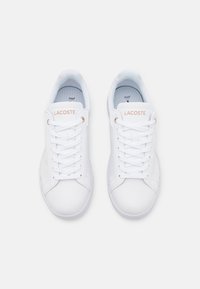 Vita sneakers i läder med en slät yta, med platta snören, rund tå och "LACOSTE"-logotyp i ljusrosa på sidan.