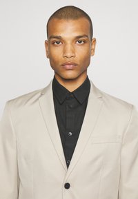Giacca di suit beige chiaro con bavero a punta, abbinata a una camicia nera con colletto. La giacca presenta un singolo bottone e una tasca sul petto.