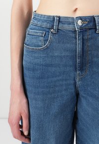 Jeans din denim de culoare bleu deschis, cu talie înaltă, cinci buzunare și detalii de cusătură subtile.Țesătura are o textură netedă și un croi clasic.