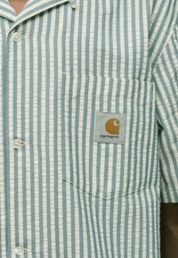 Ελαφρώς μπλε και λευκό ριγωτό πουκάμισο με κοντά μανίκια, με τσέπη στο στήθος που φέρει κεντημένο λογότυπο και κουμπιά για κλείσιμο. Μαλακό, υφή ύφασμα.