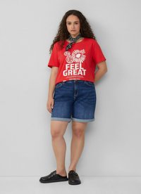 Czerwona koszulka z krótkim rękawem z białym napisem i grafiką kwiatową, w połączeniu z podwiniętymi dżinsowymi szortami i czarnymi butami typu slip-on.