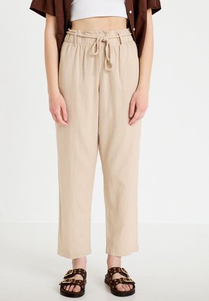 Beige high-waist paperbag-broek met koordceintuur gedragen met een wit cropped topje, een bruin open overhemd en bruine sandalen met gouden gespen.