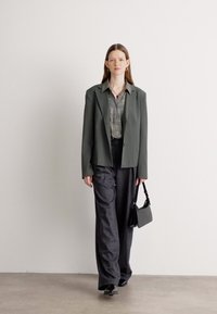 Übergrößiger grauer Blazer über einem gemusterten grün-weißen Hemd, kombiniert mit lockeren grauen Hosen, schwarzen Schuhen und einer kleinen schwarzen Handtasche.