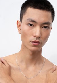 Silberkette, flaches Design, glatte Textur, die ein freies Schlüsselbein ergänzt. Das Modell hat kurze Haare und dezente Ohrstecker.