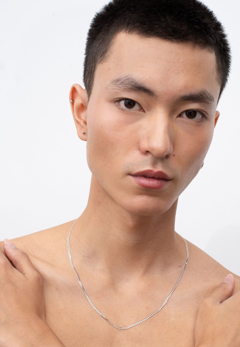 Silberkette, flaches Design, glatte Textur, die ein freies Schlüsselbein ergänzt. Das Modell hat kurze Haare und dezente Ohrstecker.