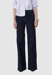 Femme portant un jean large taille haute bleu marine, une chemise blanche à boutons avec des manches bouffantes, et un bracelet en argent chunky au poignet gauche.