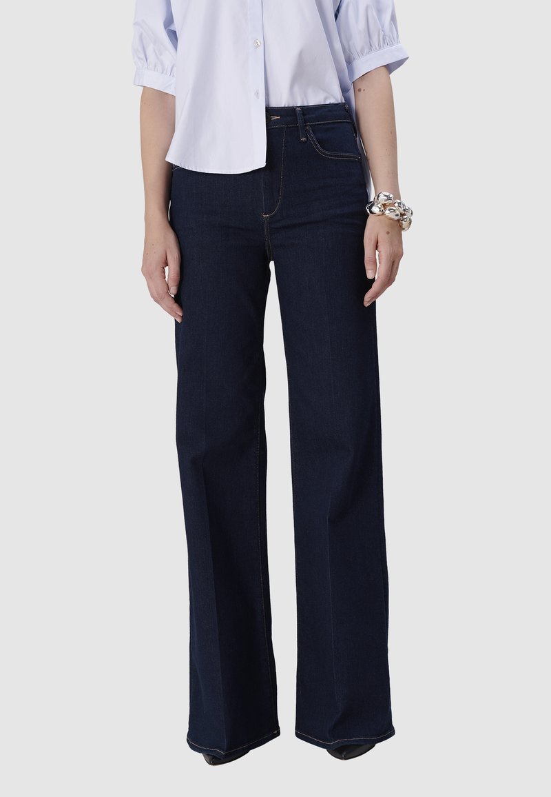 Femme portant un jean large taille haute bleu marine, une chemise blanche à boutons avec des manches bouffantes, et un bracelet en argent chunky au poignet gauche.