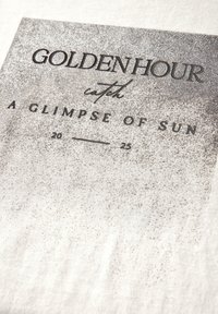 T-shirt en tissu crème clair avec un imprimé graphique comportant le texte "HORAIRE DORÉ" et "UN APERÇU DU SOLEIL" en tons gris foncé.