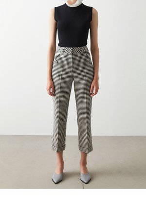 Kobieta ubrana w czarny top bez rękawów z białym marszczonym kołnierzem oraz krótkie spodnie w czarno-białą kratę z wysokim stanem, do tego pasujące spiczaste buty.