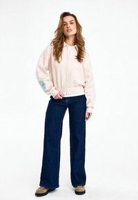 Žena stojaca v svetloružovej bunde so zipsom, tmavomodrých širokých rifle a svetlohnedých turistických topánkach na bielom pozadí.