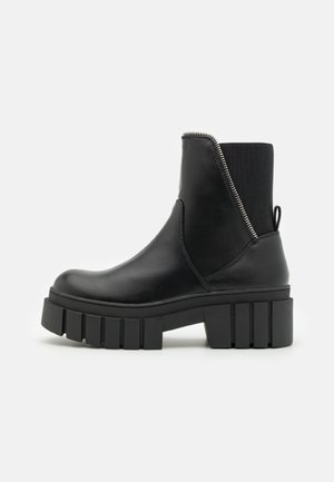 Bottine noire à semelle épaisse avec fermeture éclair latérale et panneau élastique sur fond gris neutre.