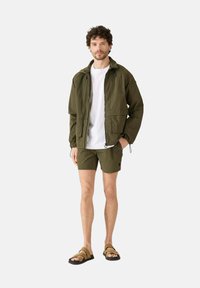 Olijfgroene lichtgewicht jas met elastische manchet, gecombineerd met bijpassende short en bruine sandalen. De outfit heeft een eenvoudige witte T-shirt eronder.