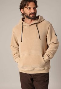 Beige hoodie gemaakt van zachte fleece met een kangoeroezak, zwarte strengen en een ronde accenten op de linker mouw.