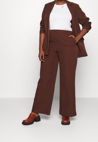 Brązowa, dopasowana marynarka w zestawieniu z białą bluzką, do tego szerokie, brązowe spodnie oraz czerwone drewniaki na grubym obcasie. Gładka tkanina o wyraźnym, starannym kroju.