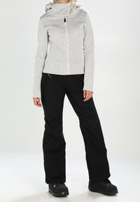 Chaqueta de cremallera gris claro con patrón texturizado, cuello alto y pantalones negros de corte holgado. Botas negras con acentos verdes.