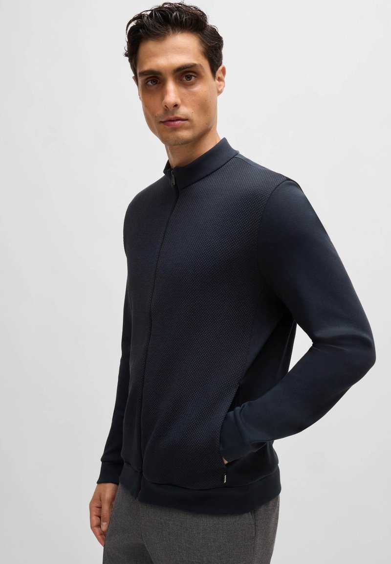 Veste zippée marine avec un tissu supérieur texturé et des manches lisses. Dotée d'un col élevé et de poches latérales. Design décontracté et ajusté.