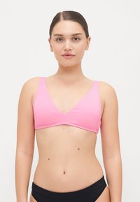 Rózsaszín bordázott bikini felsőrész V-kivágással és széles pántokkal. Simává textúrája és kontrasztos szélén kivitelezés jellemzi. A modellen fekete bikini alsó rész látható.