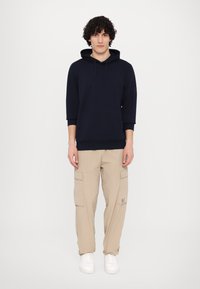 Hoodie blu navy con tasca frontale e coulisse, abbinato a pantaloni cargo beige con tasche laterali e dettaglio del logo, indossato con sneakers bianche.