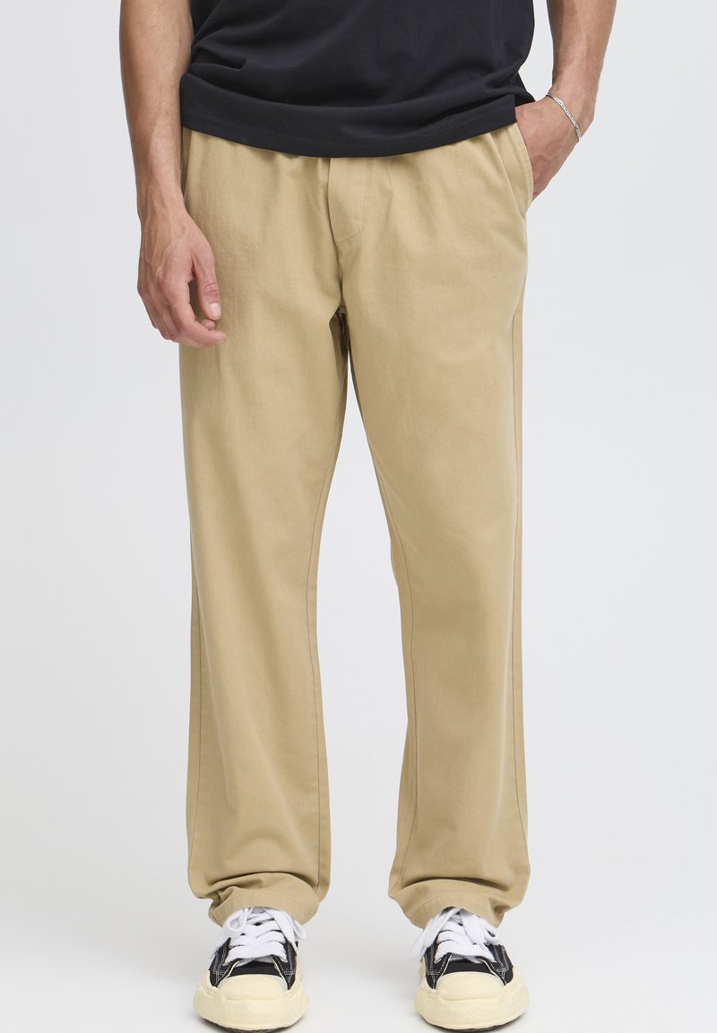 Pantalon en coton beige clair avec une coupe décontractée, doté de poches latérales et d'une coupe droite. Associé à des baskets noires et blanches.