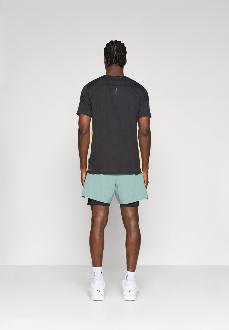 Camiseta deportiva negra con acentos laterales amarillos, combinada con pantalones cortos verde menta. Zapatillas deportivas blancas visibles, de diseño sencillo y textura suave.