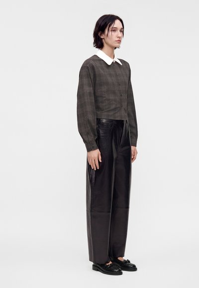 Femme debout portant une chemise à carreaux foncés boutonnée avec un col blanc, un pantalon large en cuir noir et des mocassins noirs sur fond uni.