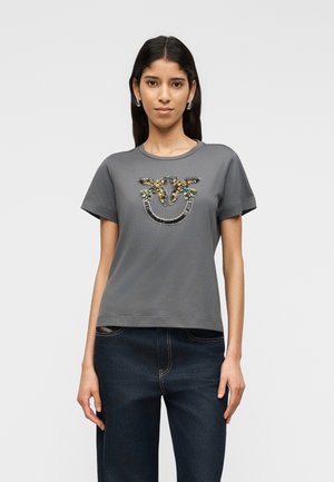 Nő szürke pólót visel, melynek mellkasán díszes két madaras mintázat található, sötétkék farmerrel, egyszínű háttér előtt áll.