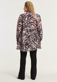 Kvinde med langt blondt hår iført en løs, langærmet bluse med pink og sort tigerstriber og sorte bukser med vidde, stående med ryggen til.