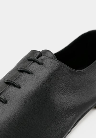 Μαύρο δερμάτινο παπούτσι με λεία υφή, με χαμηλή γλώσσα και διακοσμητικό κορδόνι στο επάνω μέρος. Χωρίς ορατά μεταλλικά στοιχεία ή διακοσμήσεις.