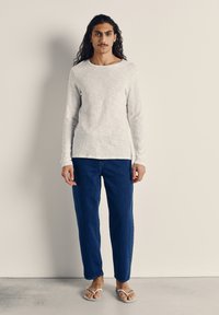 Biele tričko s dlhým rukávom a okrúhlym výstrihom, modré rifle s voľným strihom a biele žabky. Model stojí na jednoduchom pozadí.