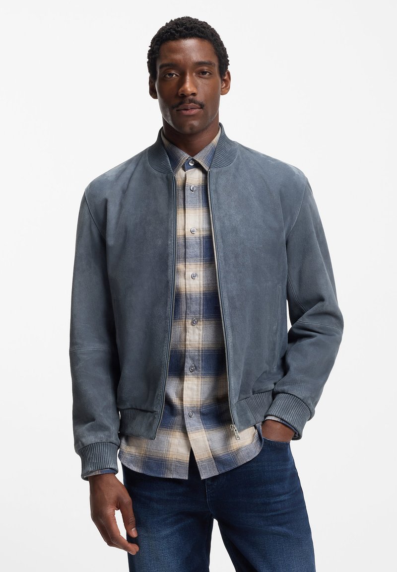 Giacca bomber in camoscio grigio scuro con polsini e colletto a coste. Indossata sopra una camicia a quadri blu e beige. Presenta una chiusura con cerniera e tasche.