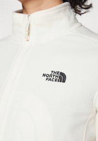 Valge fliisjakke kõrge kraega, keskel tõmblukk ja must broderitud logo vasakul rinnal. Pehme tekstuur, minimalistlik disain.