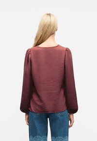 Ξανθός άνθρωπος που φαίνεται από πίσω, φοράει ένα μπορντό μπλούζα μακρυ μάνικη με υφή και μπλε τζιν με δαντελωτά σχέδια στα τελειώματα.