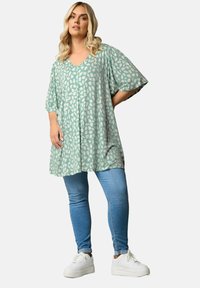 Mintgrön tunika med V-ringning, korta ärmar och ett vitt bladmönster, i kombination med ljusblå jeans och vita sneakers.