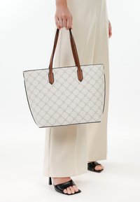 Borsa tote bianca con un motivo geometrico, bordatura nera e manici in pelle marrone, tenuta da una persona che indossa pantaloni beige chiaro.