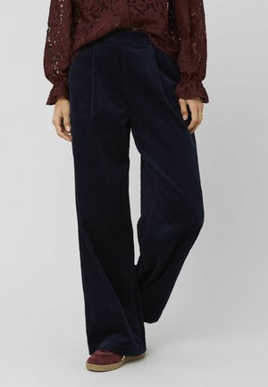 Pantalons larges en velours côtelé bleu marine avec des rayures verticales, deux plis avant et une taille lisse, associés à des chaussures décontractées.