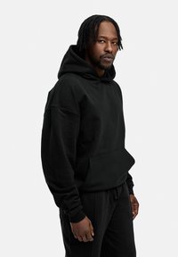 Schwarzer Kapuzenpullover aus weichem Material. Verfügt über eine Fronttasche, eine lässige Passform und gerippte Bündchen. Kombiniert mit passenden schwarzen Jogginghosen.