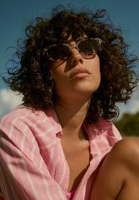 Des lunettes de soleil en œil de chat avec un motif écaille de tortue, portées avec une chemise rayée rose. Les cheveux naturels et bouclés sont coiffés de manière lâche.