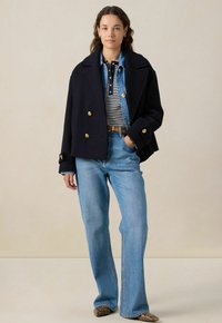 Manteau marin double boutonnage bleu marine avec des boutons dorés, superposé à une chemise rayée, associé à un jean bleu à jambe large et des chaussures pointues à motif serpent.