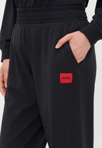 Zwarte joggingbroek van zacht materiaal, met een elastische tailleband, zijzak en een rode label met het "HUGO" logo aan de linkerkant.