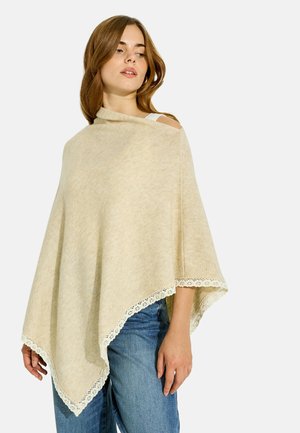 Mujer con un poncho de punto beige con ribete de encaje sobre una camiseta blanca y vaqueros azules, mirando ligeramente hacia un lado sobre un fondo sencillo.