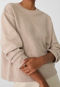 Person som bär en beige stickad tröja med upprullade ärmar, håller ett handled med den andra handen, bär en guld ring med en svart sten.