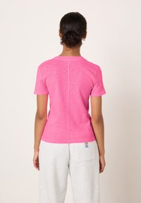 Persona con capelli ricci raccolti in uno chignon, che indossa una maglietta aderente rosa a maniche corte e pantaloni grigio chiaro, in piedi con la schiena rivolta verso la fotocamera.